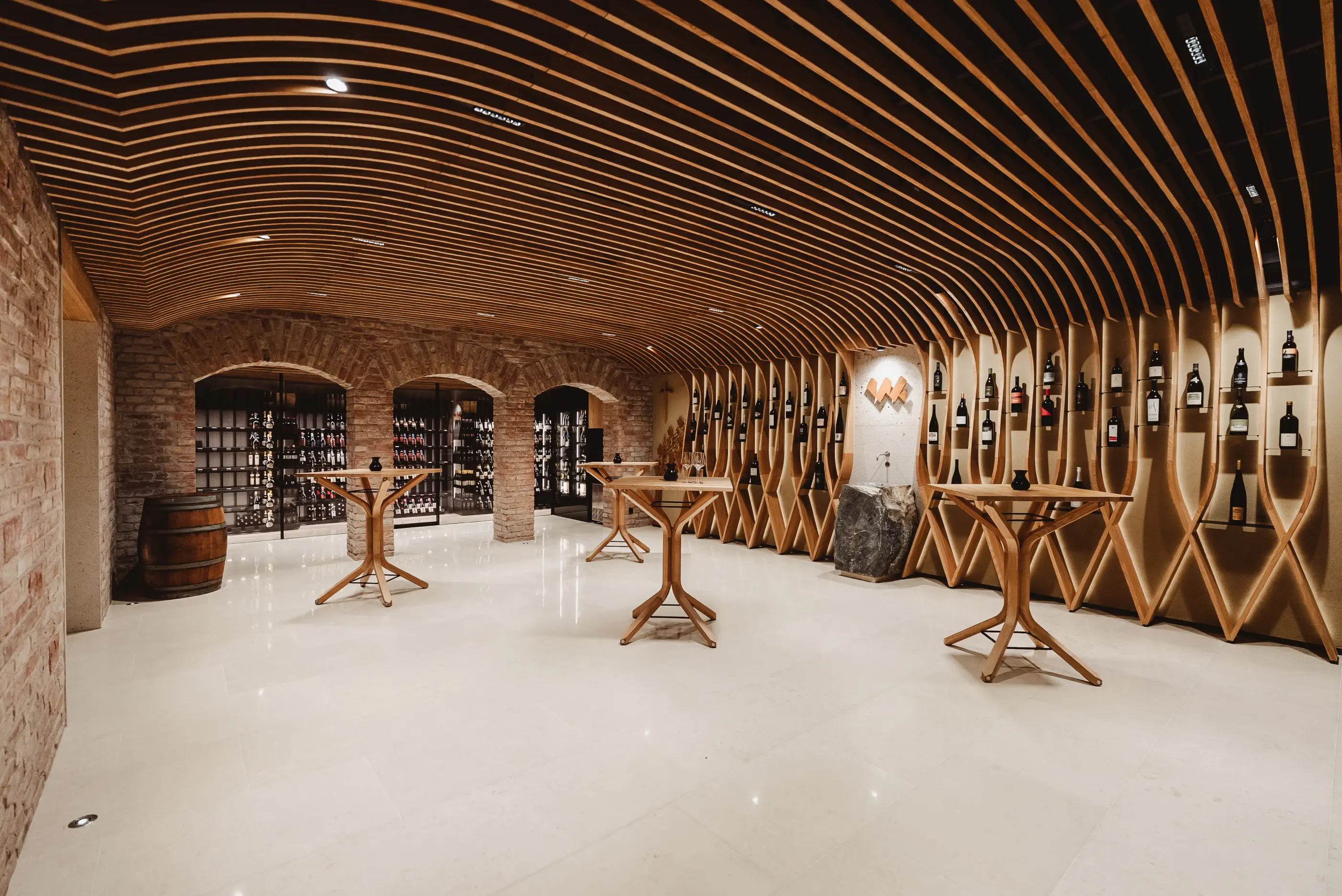 Modern wine cellar with wooden curved ceiling, wine racks, brick walls, and standing tables.