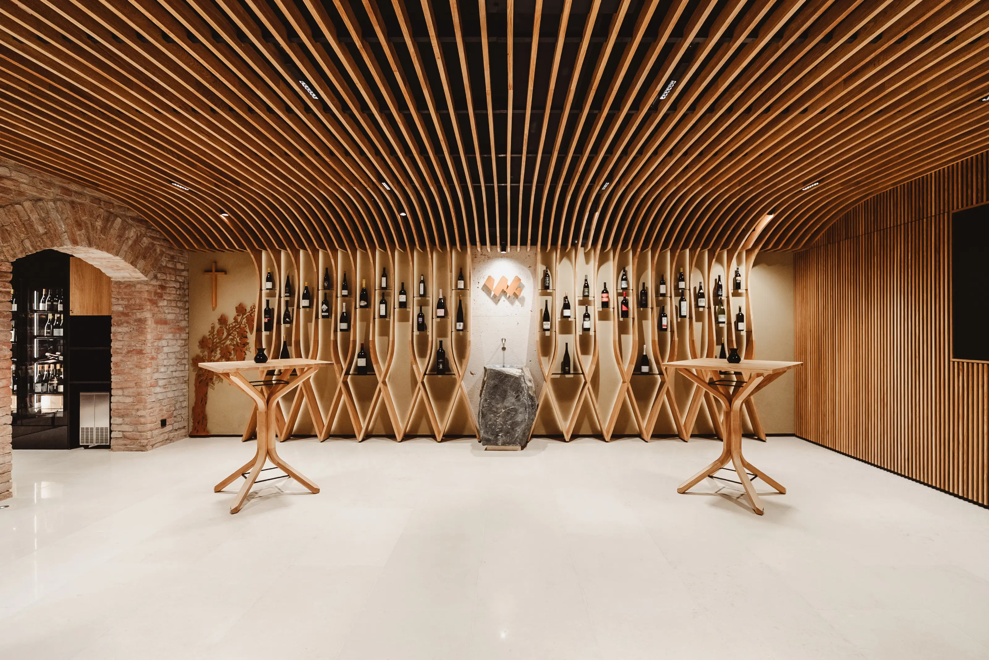Modern wine cellar with wooden curved ceiling, sculptural wine racks, stone pedestal, and two wooden tables.