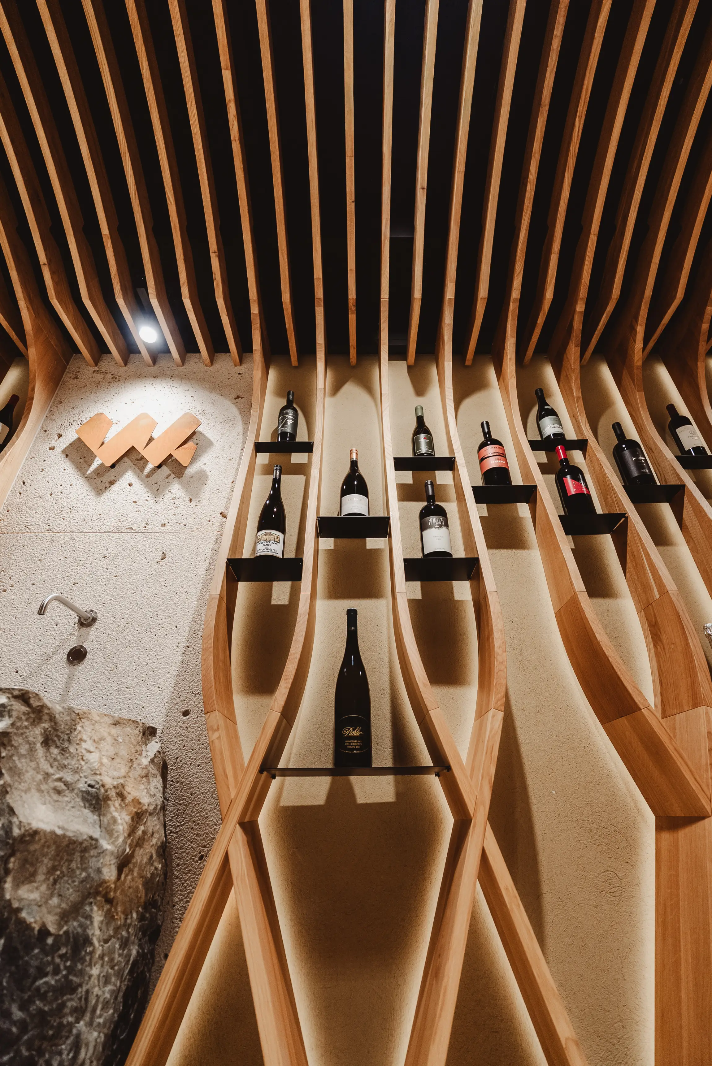 Modern wooden wine rack with various wine bottles displayed vertically on shelves against a textured wall.