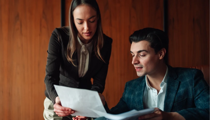A man and woman in business attire review documents together in an office with wood panel walls.