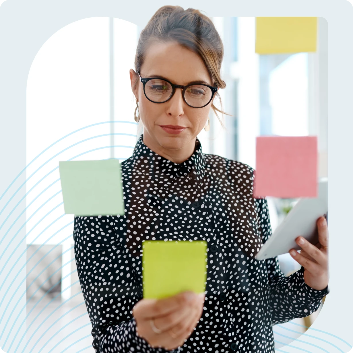 Woman in glasses placing colorful sticky notes on a glass wall while holding a tablet.