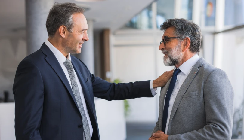 Two professional men in suits smiling and talking in an office, one with hand on the other's shoulder.