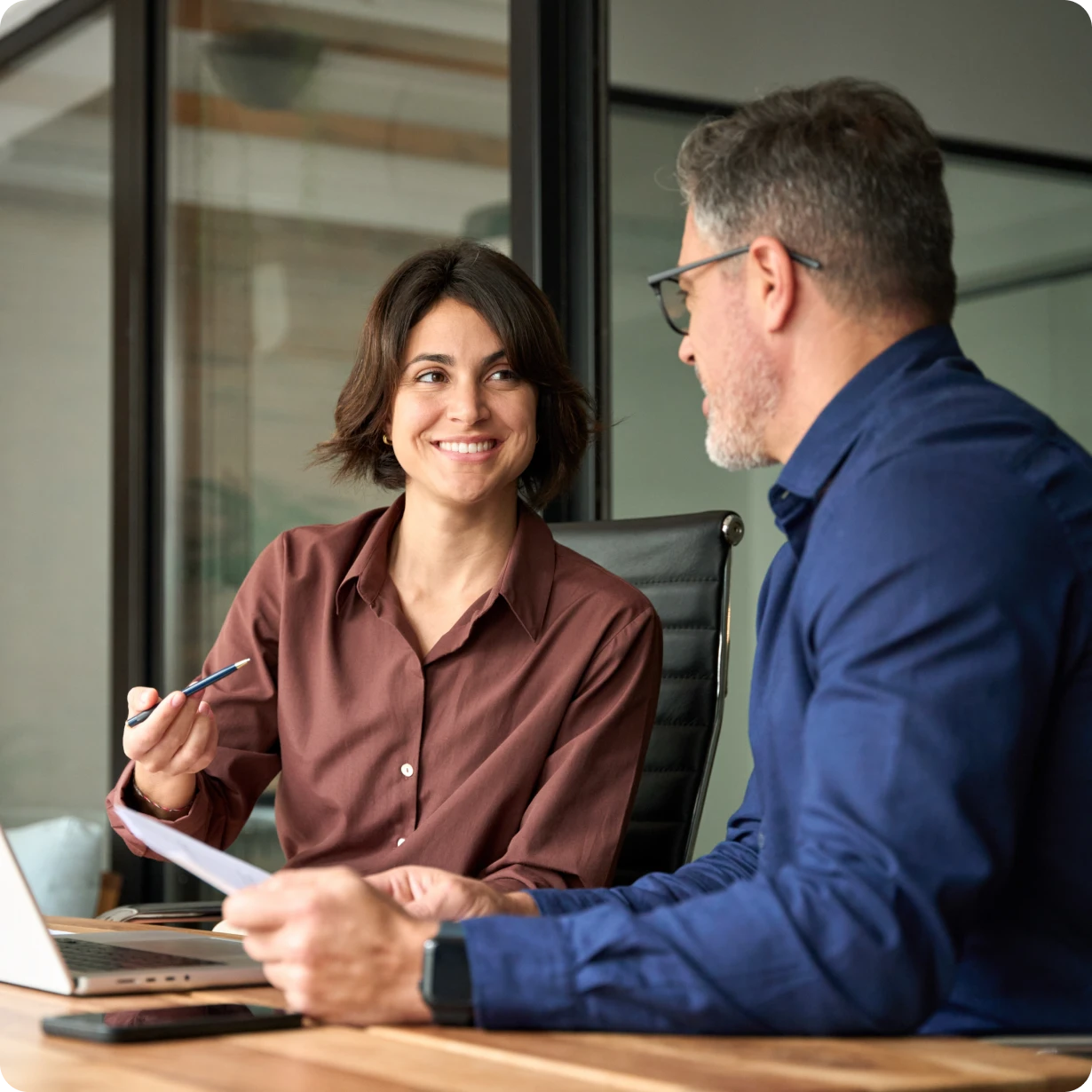 Woman smiling and holding a pen while talking to a man in a blue shirt at a desk with a laptop.