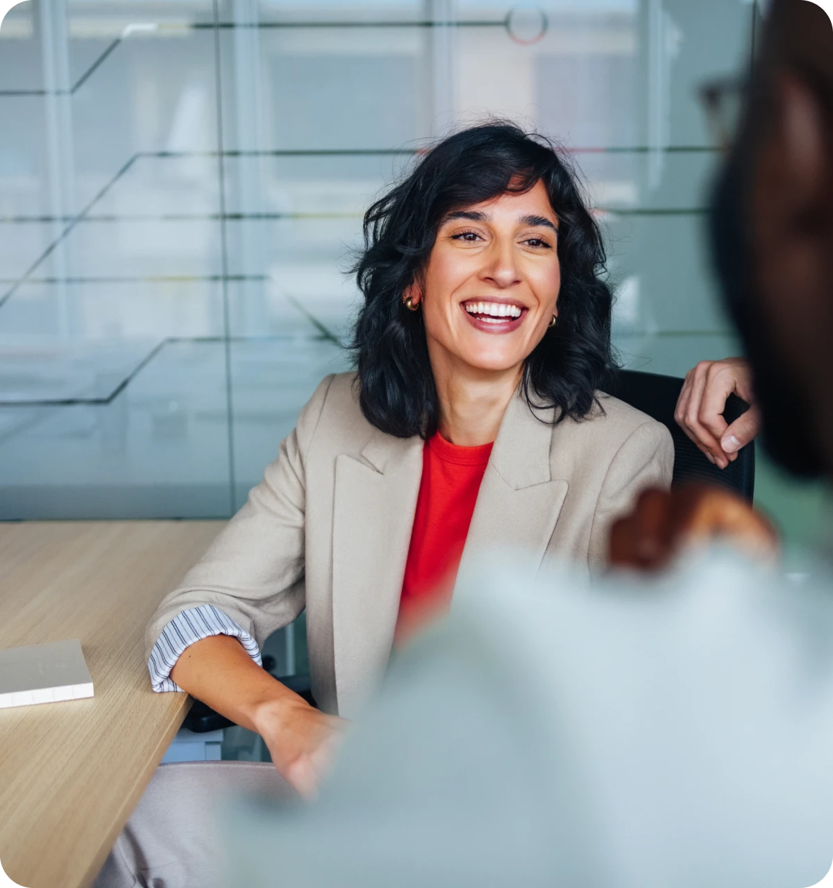 Smiling woman in beige blazer and red top sitting and talking in modern office meeting room.