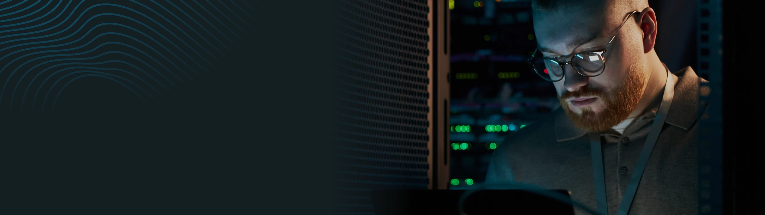 Man with glasses working on a laptop inside a dimly lit server room with glowing equipment.