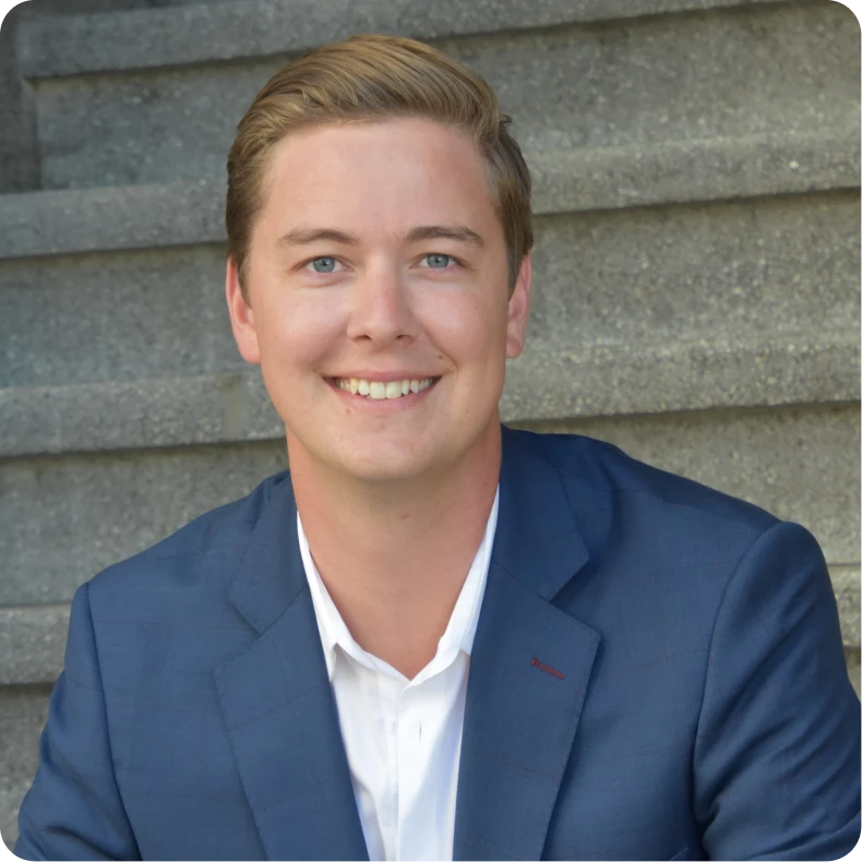 Smiling young man with blond hair wearing a blue suit jacket and white shirt sitting on concrete steps.
