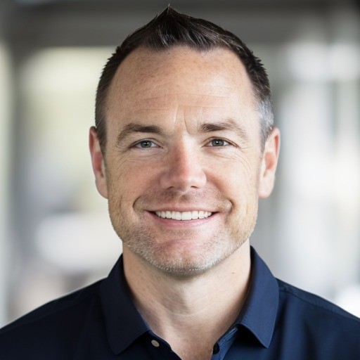 Smiling man with short dark hair wearing a navy blue collared shirt against a blurred background.