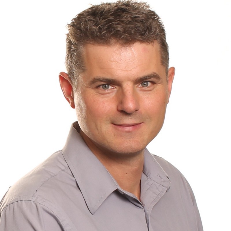 Man with short curly hair wearing a gray collared shirt smiling against a white background.
