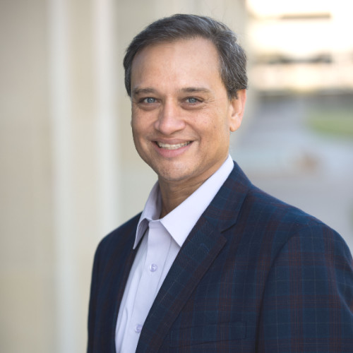 Smiling man in a dark plaid blazer and light shirt standing outdoors near a building.