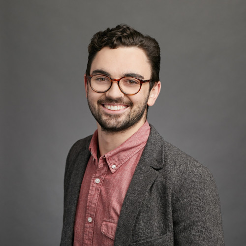 Smiling man with glasses wearing a gray blazer over a red shirt against a gray background.