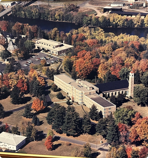 Aerial view of large buildings surrounded by autumn trees with colorful foliage near a riverbank.