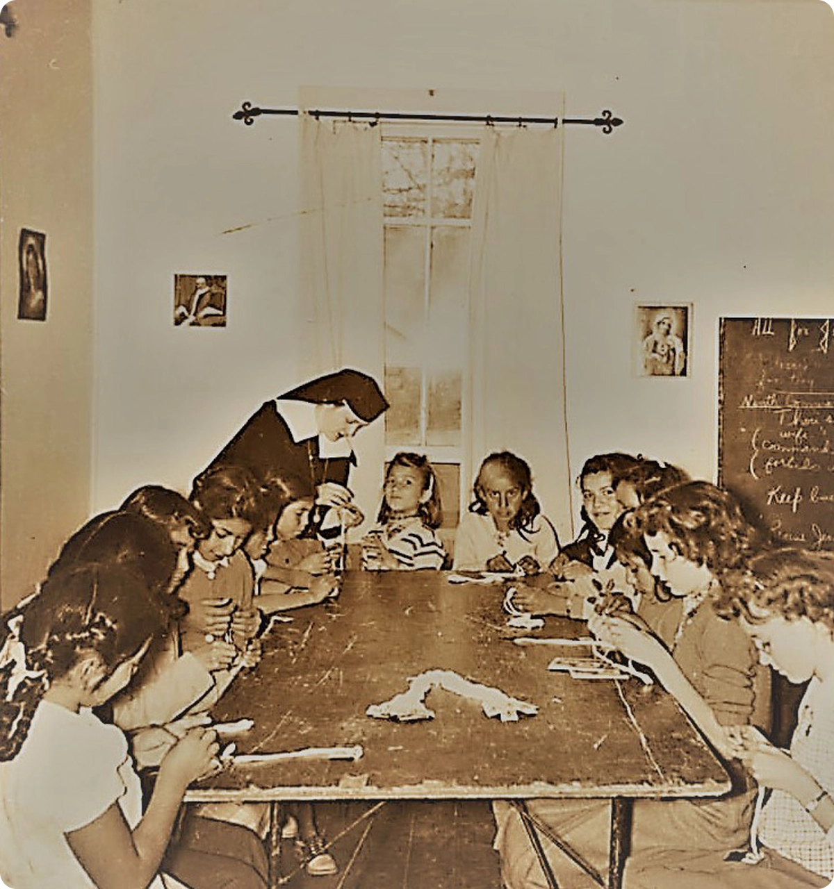 Sepia-toned photo of a nun teaching a group of girls seated around a wooden table, working with yarn or thread.