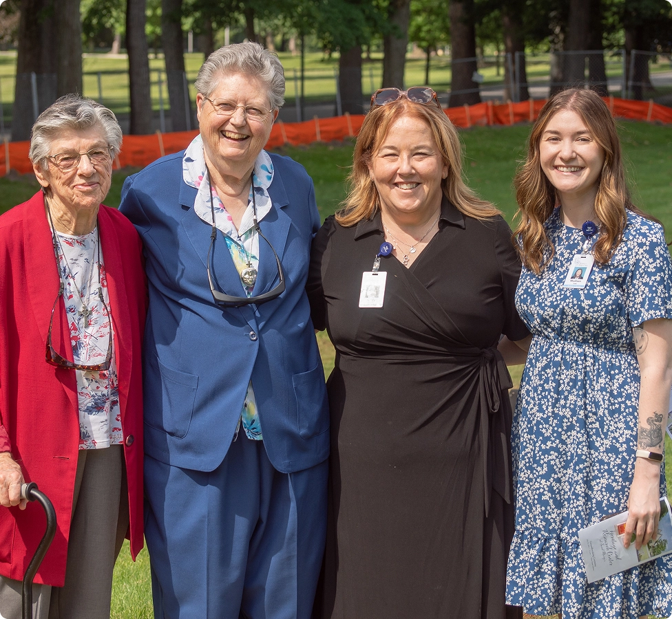 Four smiling women standing outdoors on grass with trees in the background, two elderly and two younger adults.
