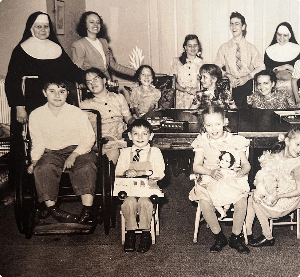 Black and white photo of children and nuns in a classroom, some children hold toys and dolls.