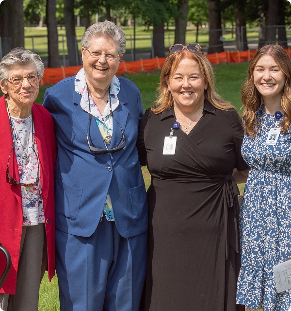 Four women smiling outdoors with trees and grass in the background.