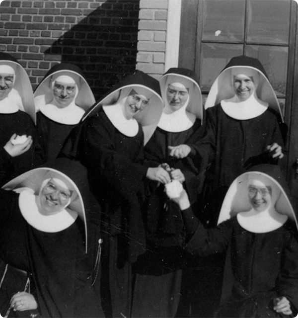 Group of seven smiling nuns in traditional habits standing and sitting outside a brick building.