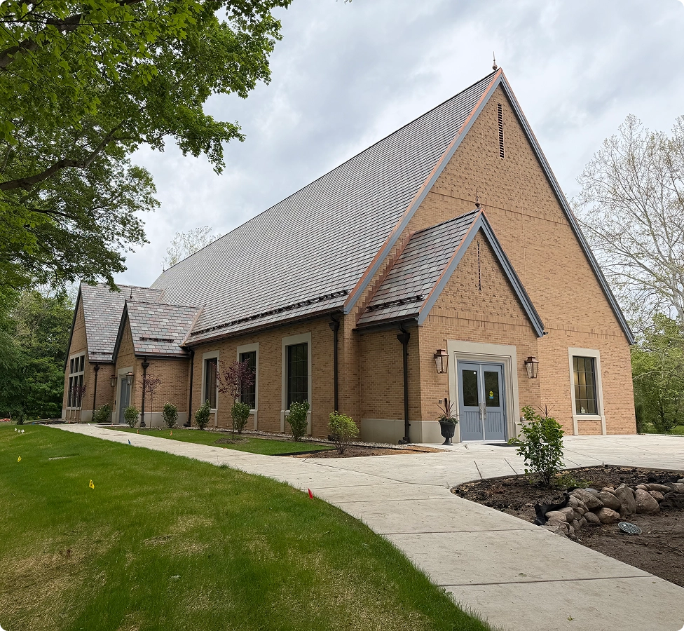 Large brick building with steep slate roof, blue double doors, and surrounding sidewalk and greenery.