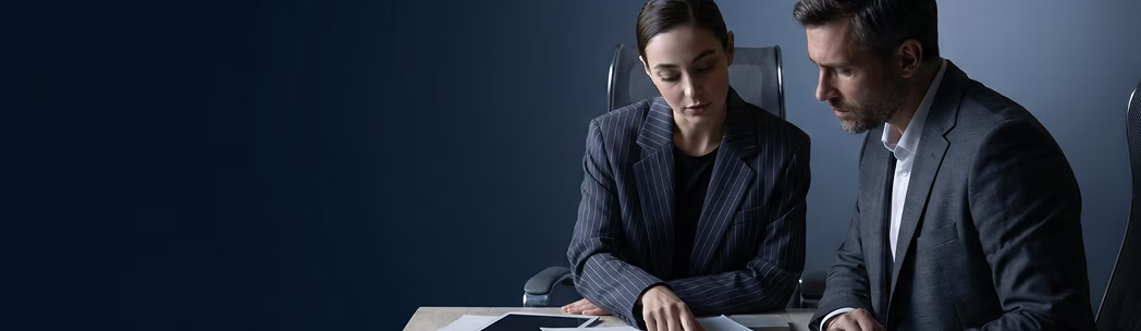 Two business professionals reviewing documents together at a desk against a dark blue background.