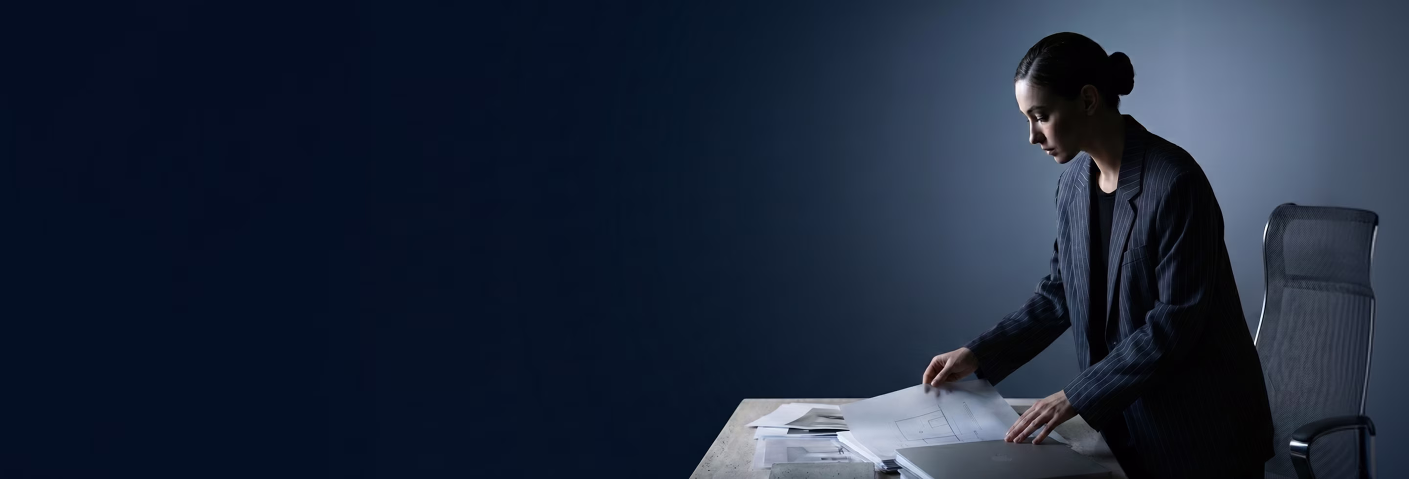 Woman in a pinstripe blazer reviewing architectural drawings at a desk against a dark background.