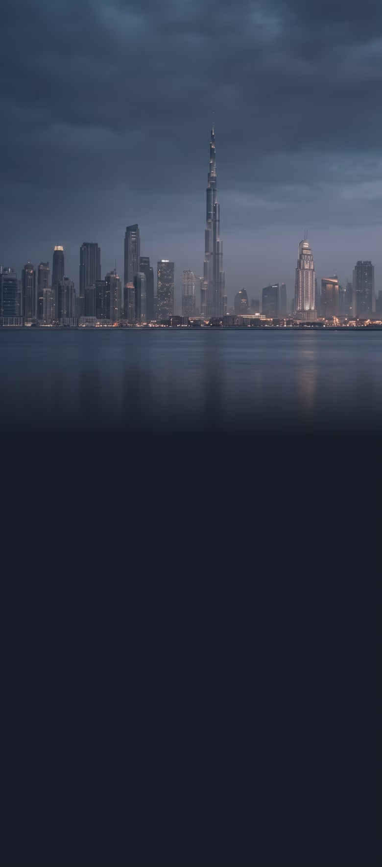 Night skyline of a city with tall illuminated skyscrapers reflected on calm water under a cloudy sky.