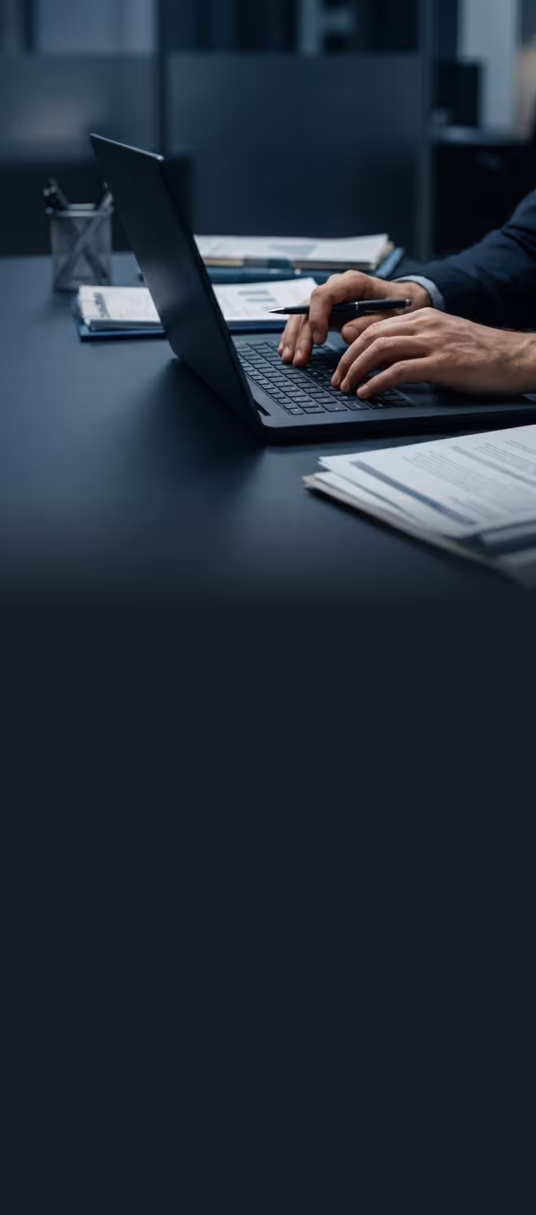 Person typing on a laptop with documents and a pen on a dark office desk.