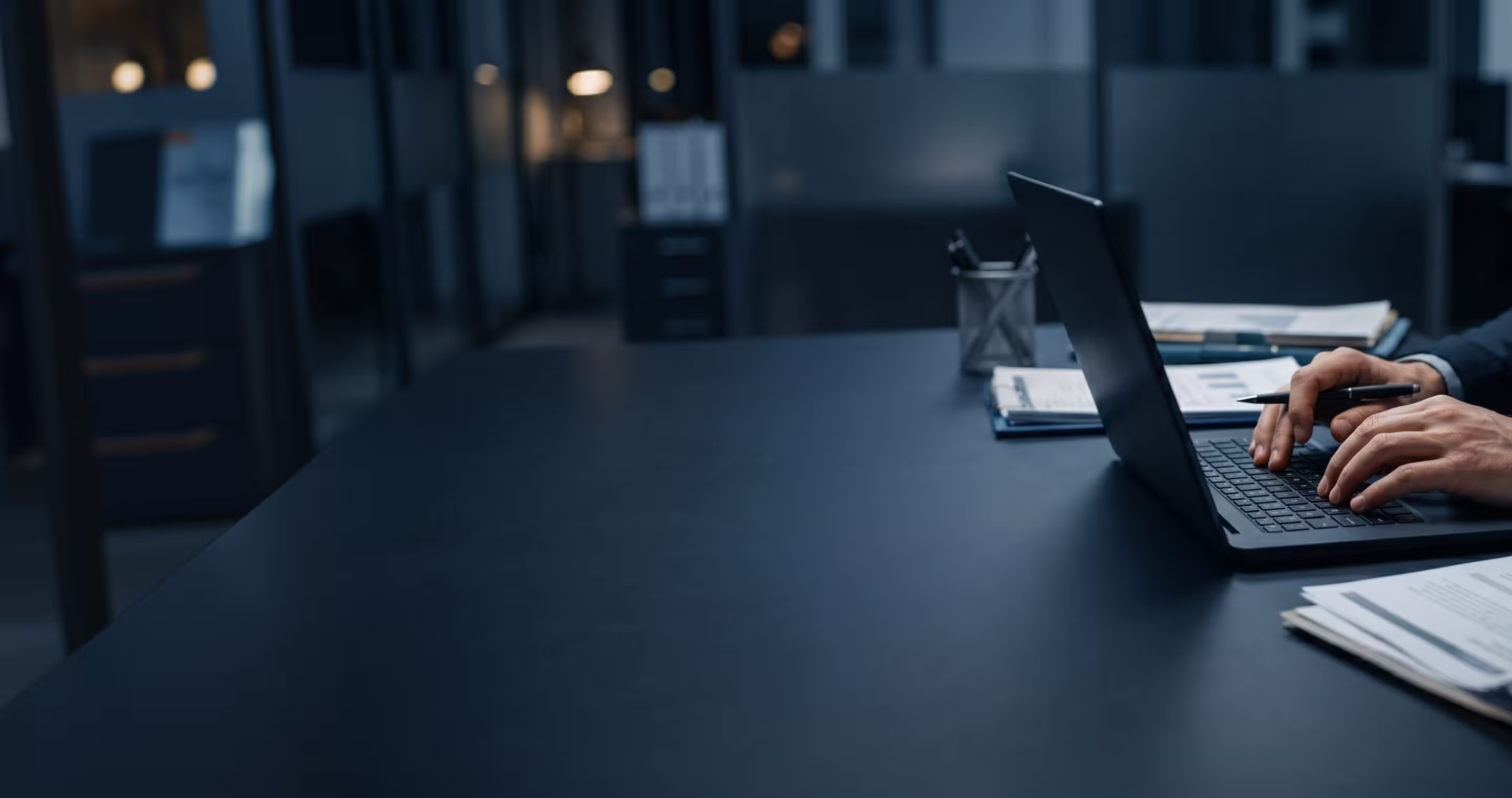 Person typing on a laptop with documents and pen on a dark office desk.