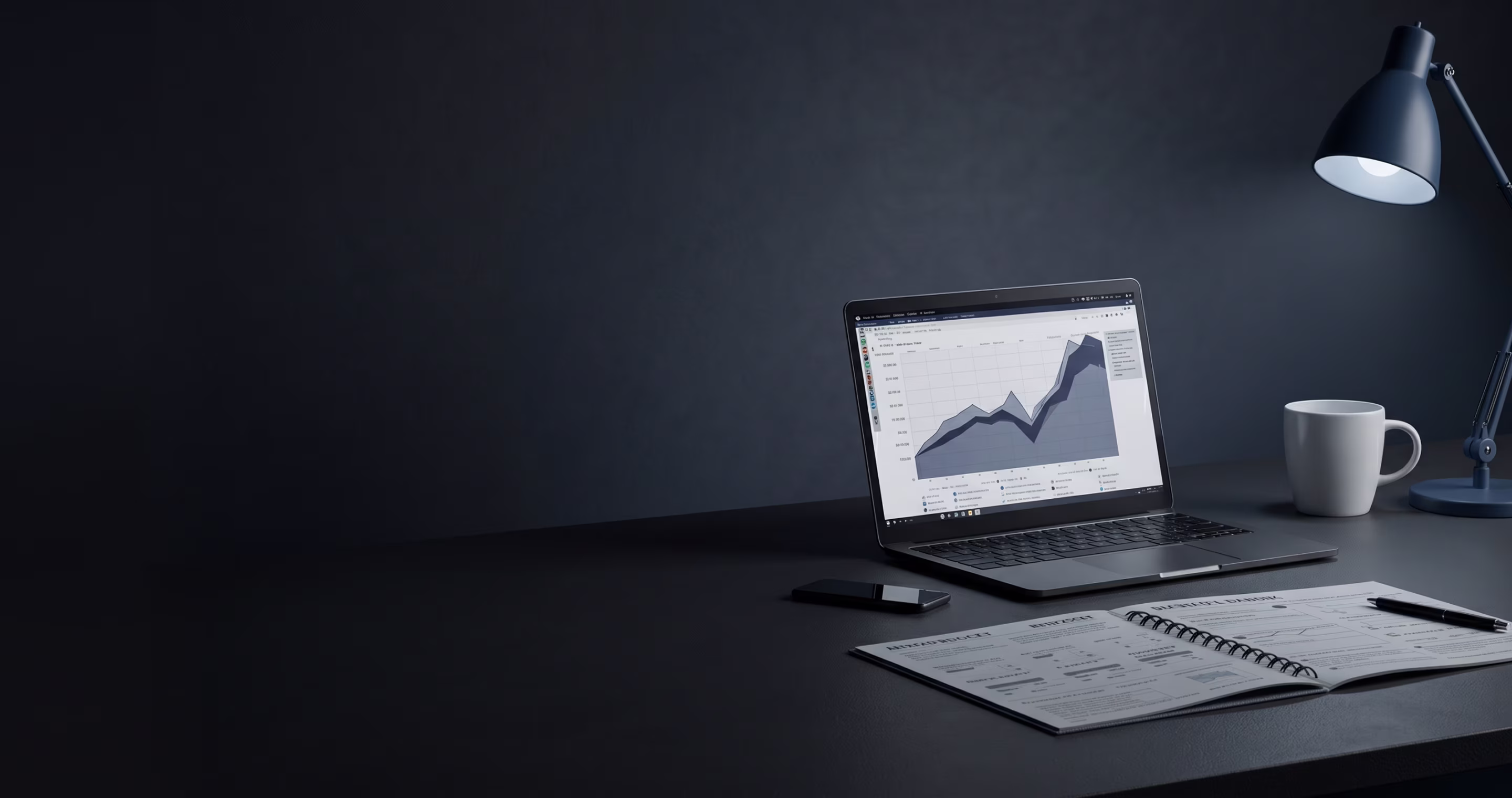 Laptop displaying a graph on a dark desk with a smartphone, open notebook with pen, lamp, and white mug.