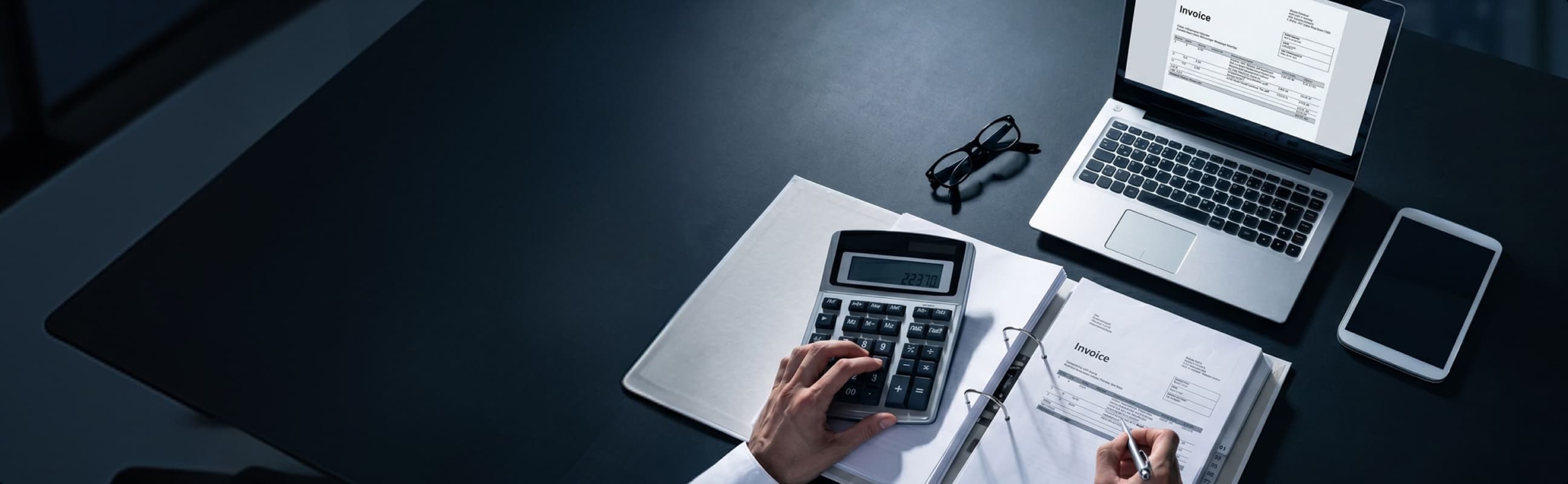 Person calculating invoice numbers on a calculator with a laptop, tablet, and glasses on a dark desk.