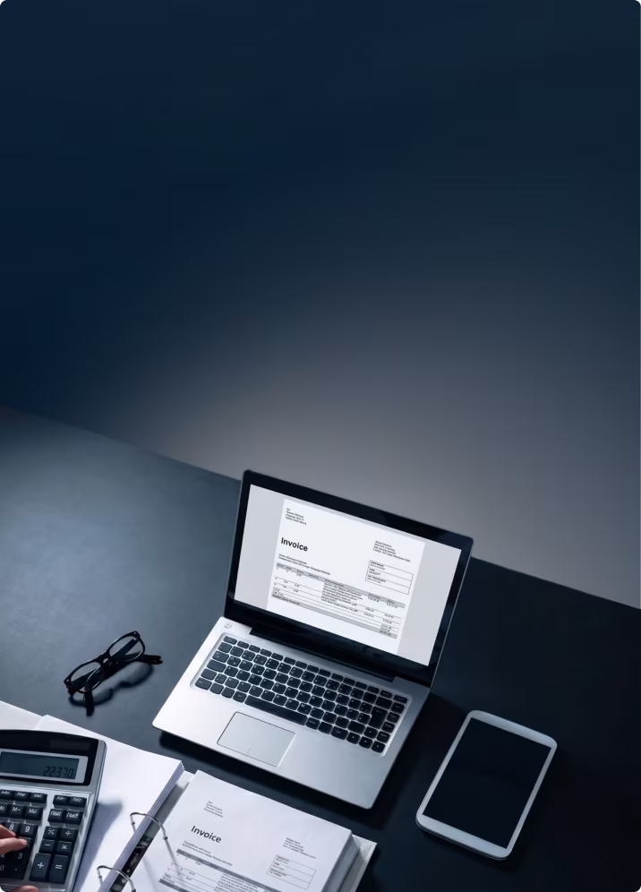 Laptop displaying an invoice on screen with a calculator, printed invoices, glasses, and a tablet on a dark desk.