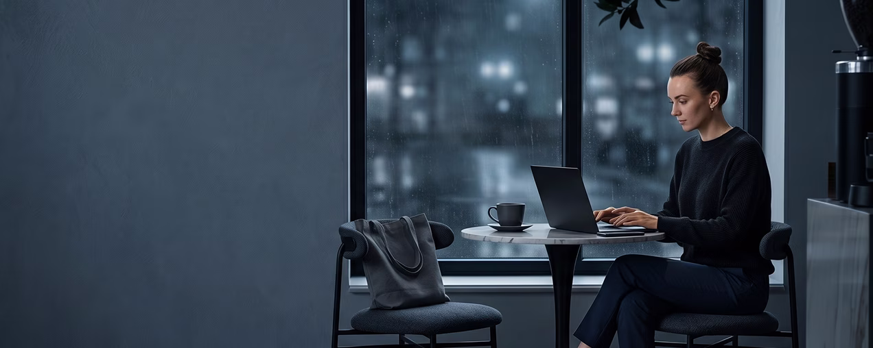 Woman in dark clothes typing on laptop at table by window with coffee cup on a rainy evening.