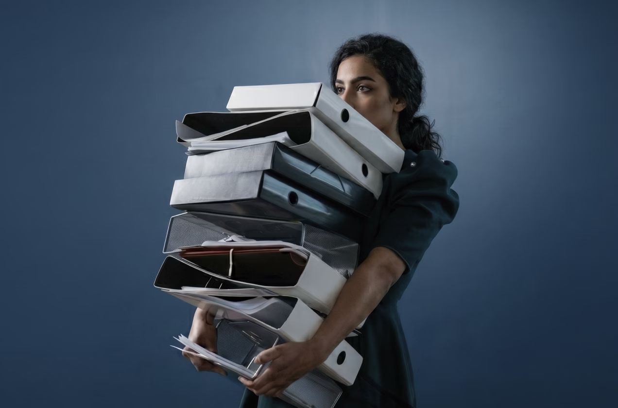 Woman holding a large stack of binders and documents against a blue background.