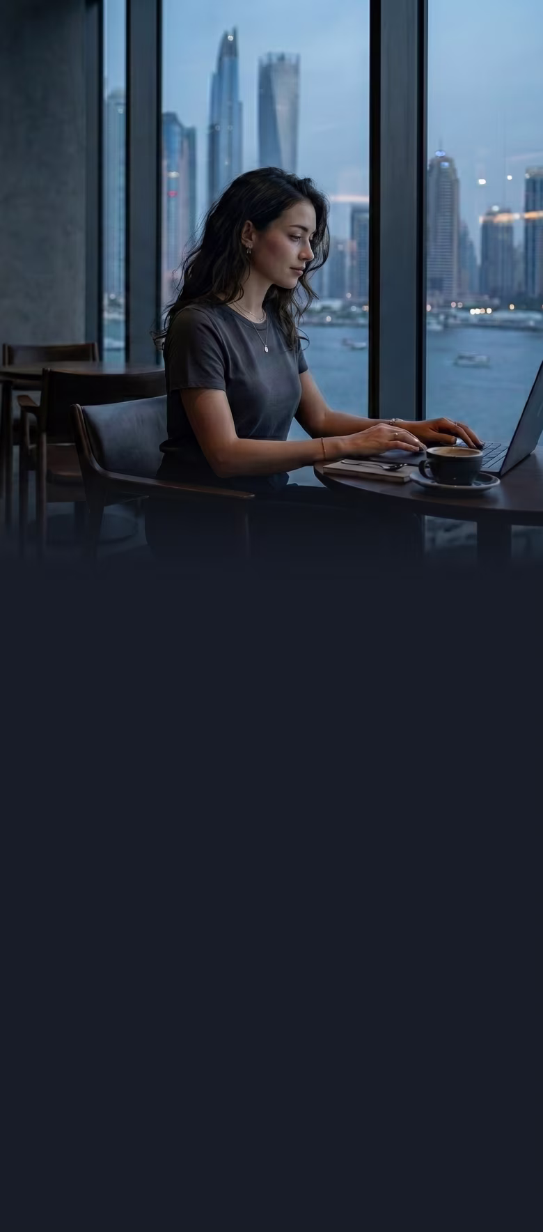 Woman working on a laptop in a cafe with a city skyline and water view through large windows.