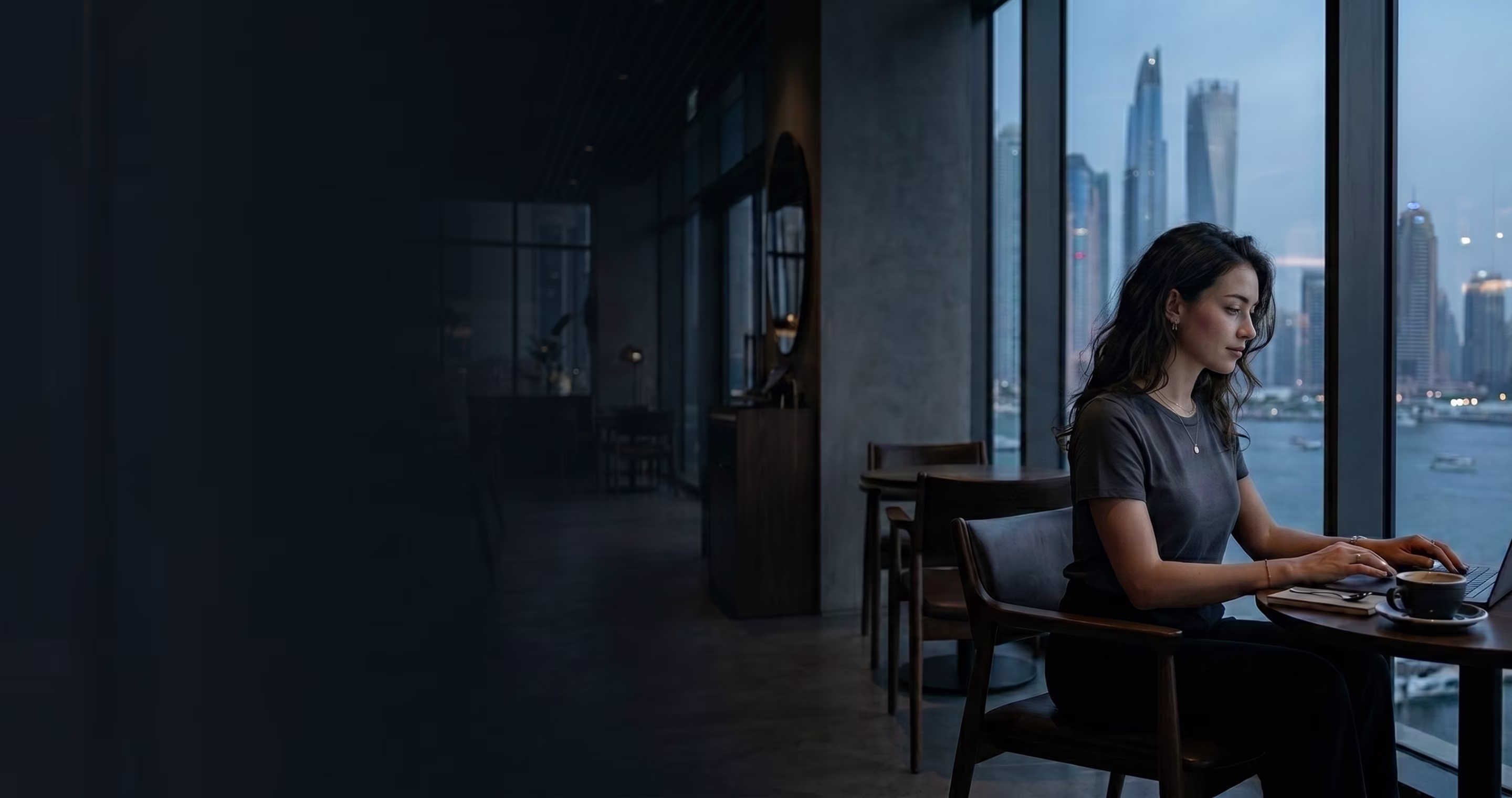 Woman working on laptop in a cafe with large windows overlooking a city skyline and river.