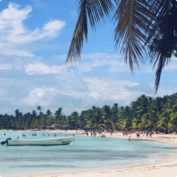 Tropical beach with palm trees, clear blue water, people swimming, and a small boat near shore.