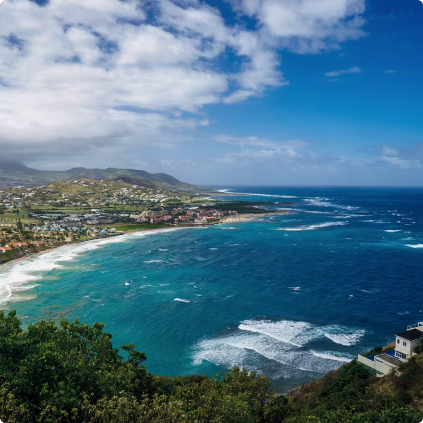 Coastal view of a tropical island with blue ocean, white waves, hills, and scattered houses under a partly cloudy sky.