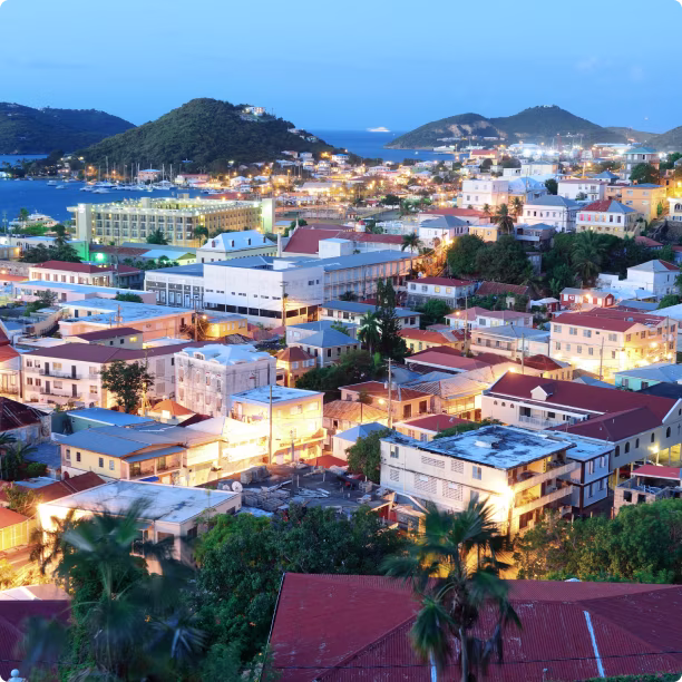 Cityscape of Antigua and Barbuda at dusk with illuminated buildings, hills, and ocean in the background.