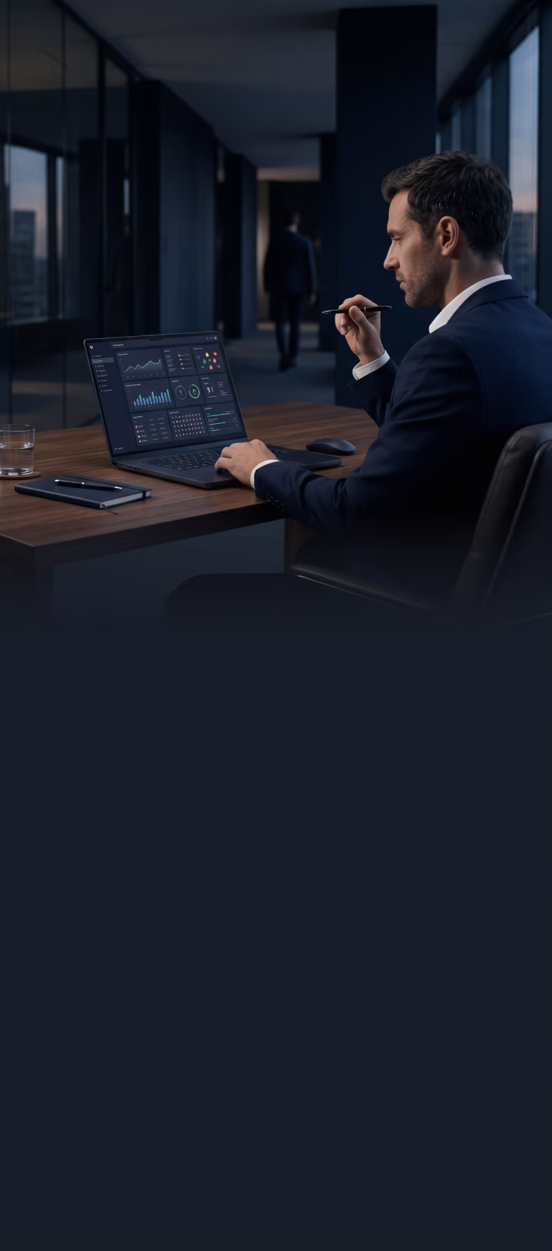 Man in suit working on laptop with data dashboard on screen in modern office at dusk.