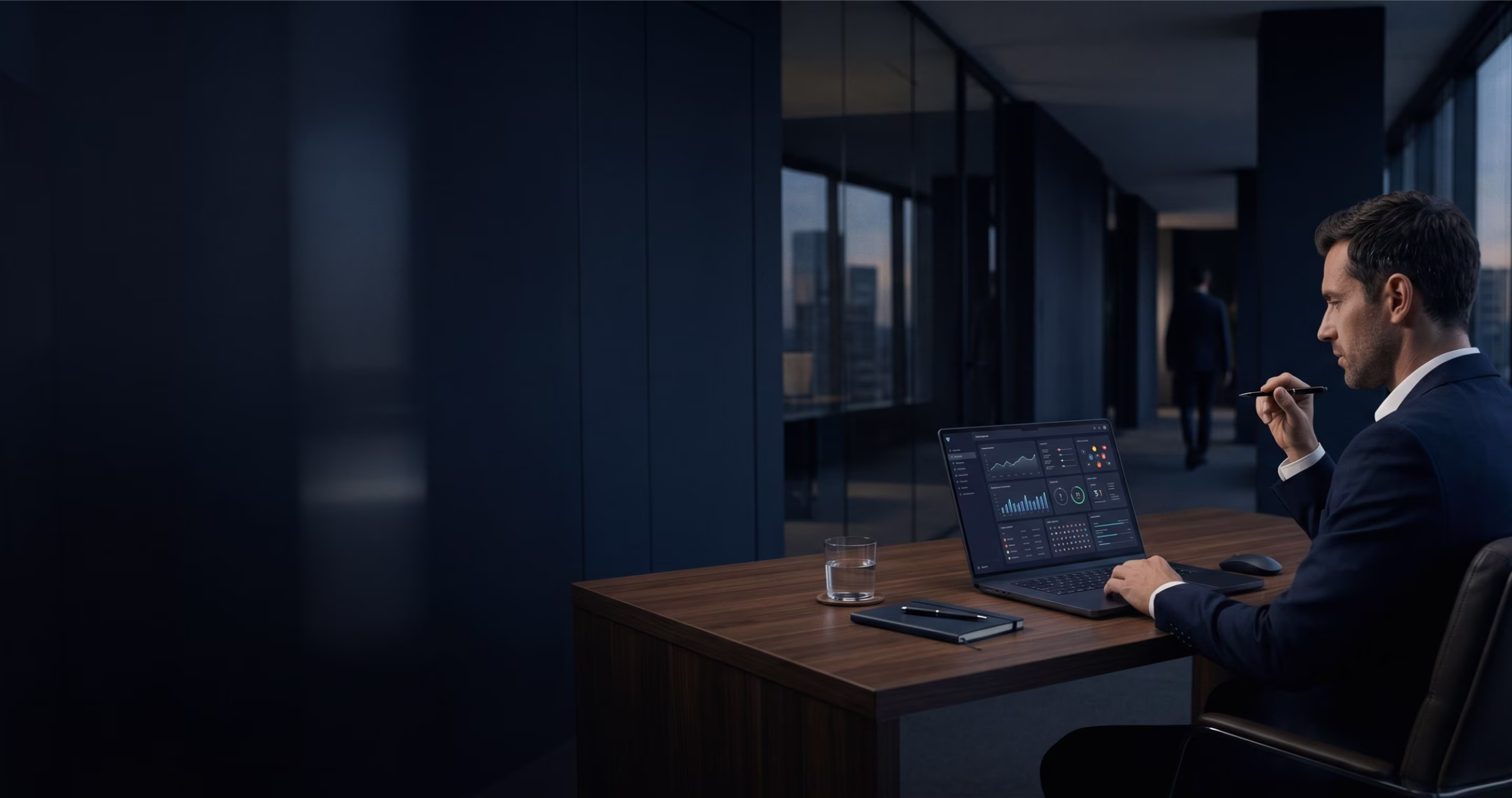 Man in suit working on a laptop with data graphs in a modern office at dusk.