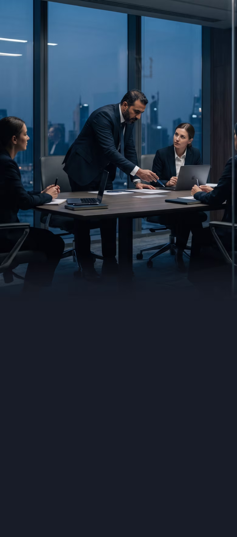 Businessman standing and pointing at documents while colleagues in suits listen in conference room.