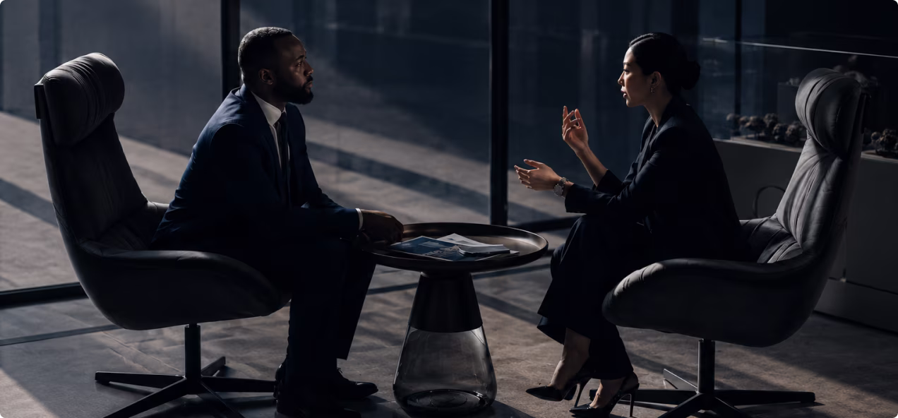 Businessman and businesswoman in suits having a discussion while seated in office chairs at a round coffee table.