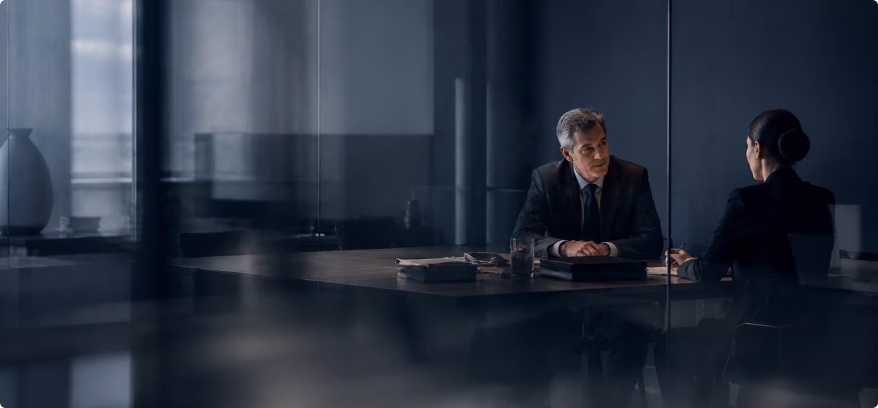 Two business professionals in dark suits having a serious discussion at a conference table in an office.