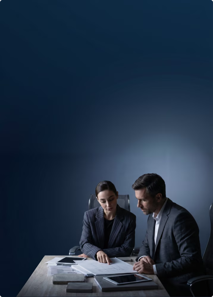 Two business professionals in suits reviewing documents together at a desk with tablets and paper.