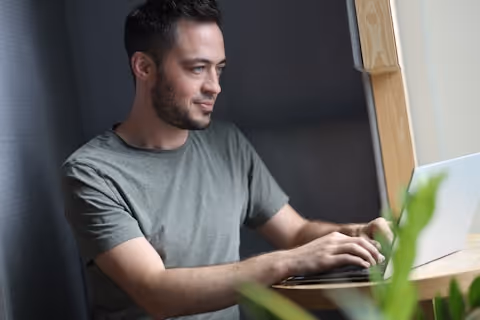 Mann mit kurzem dunklem Haar sitzt an einem hellen Fensterplatz und arbeitet an einem Laptop.