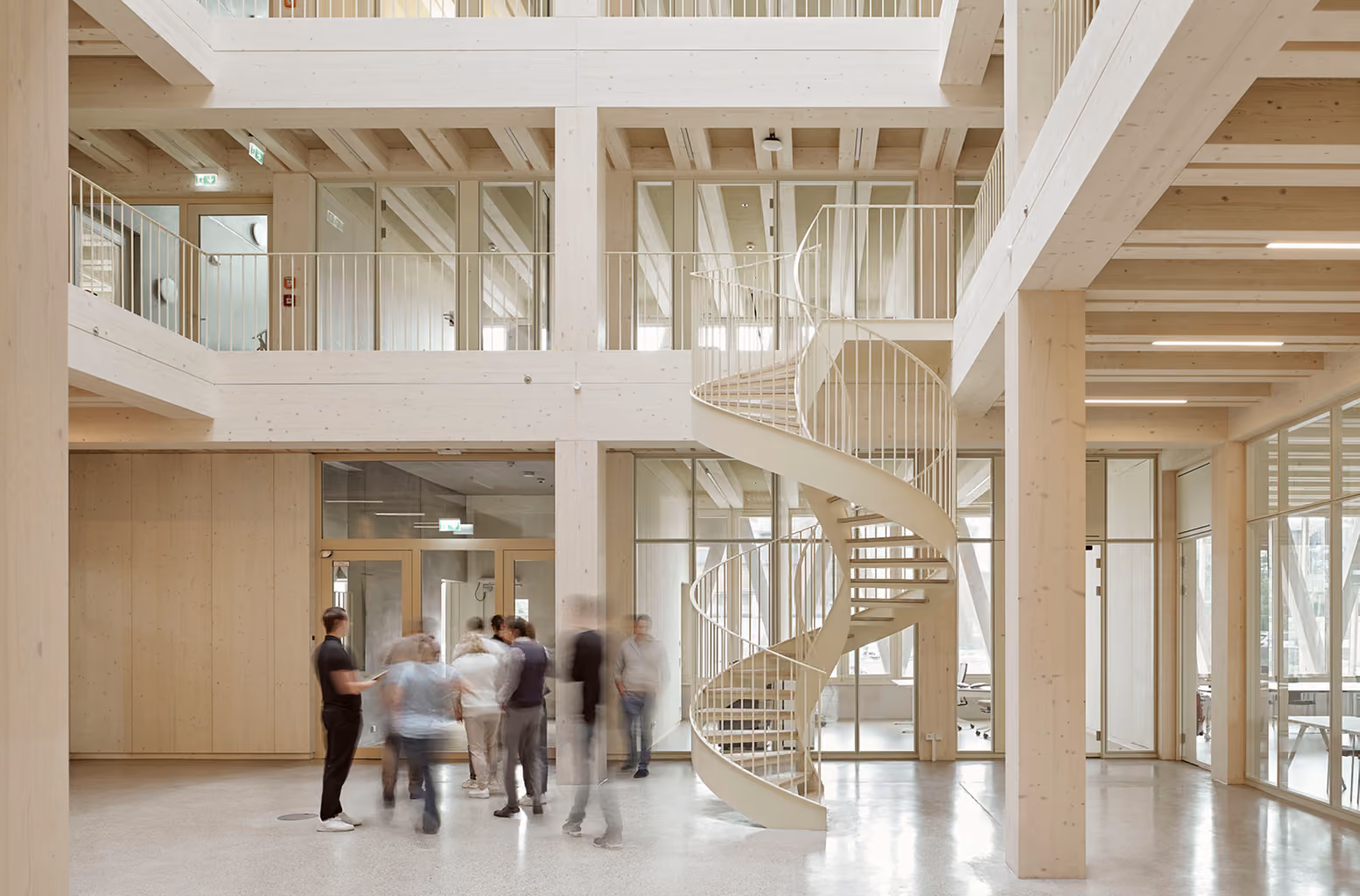 Menschengruppe im Foyer eines Holzbaus, in der Mitte eine helle Wendeltreppe über zwei Etagen.