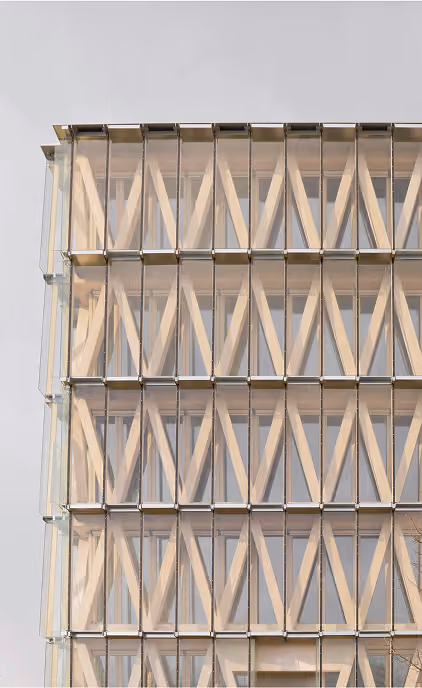 Fassade eines modernen Gebäudes mit rautenförmigem Holztragwerk hinter einer Glashülle.