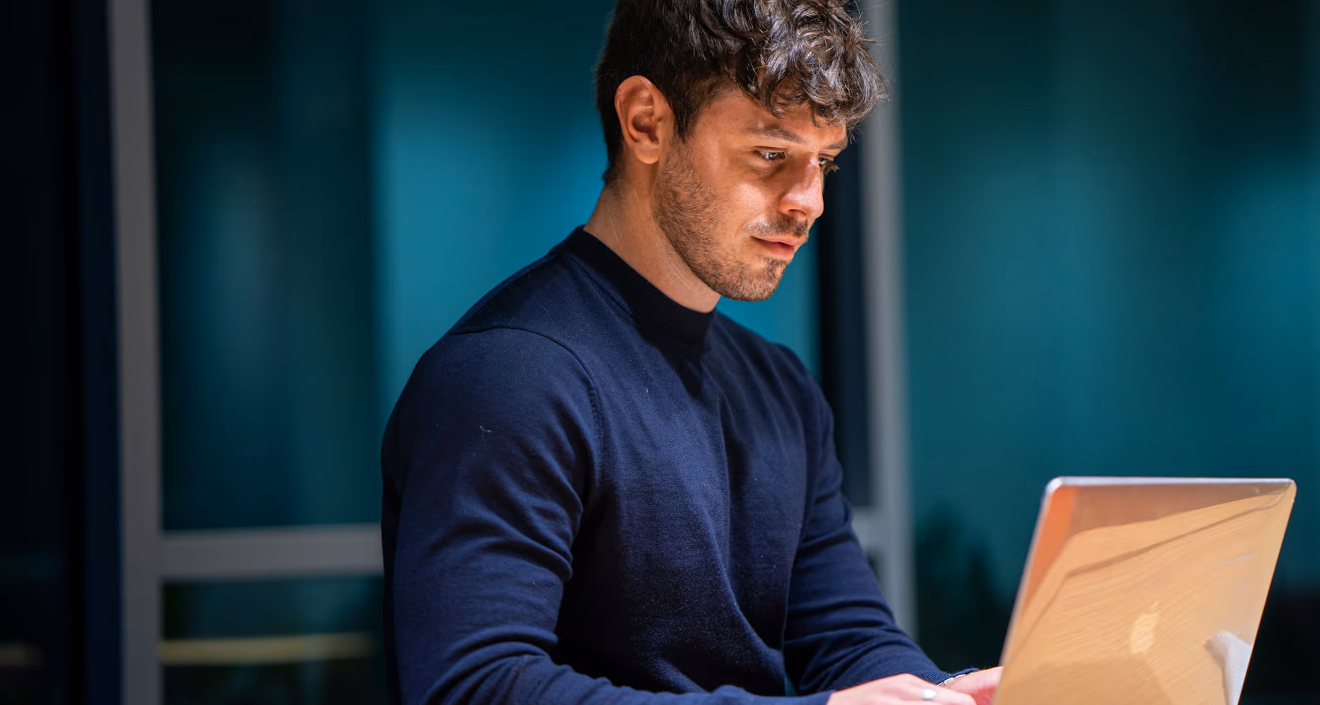 Junger Mann mit dunklem Pullover arbeitet konzentriert an einem Laptop in einem Büro mit gedämpftem Hintergrundlicht.