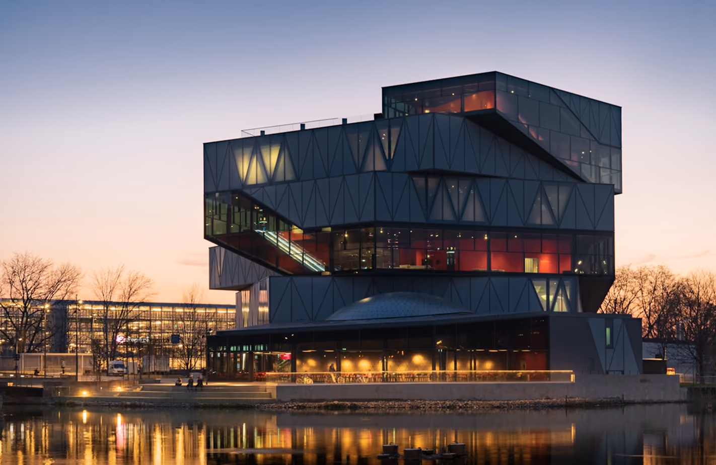 Modernes, mehrstöckiges Gebäude mit geometrischem Design und vielen beleuchteten Fenstern am Wasser bei Dämmerung, Spiegelung im See sichtbar.