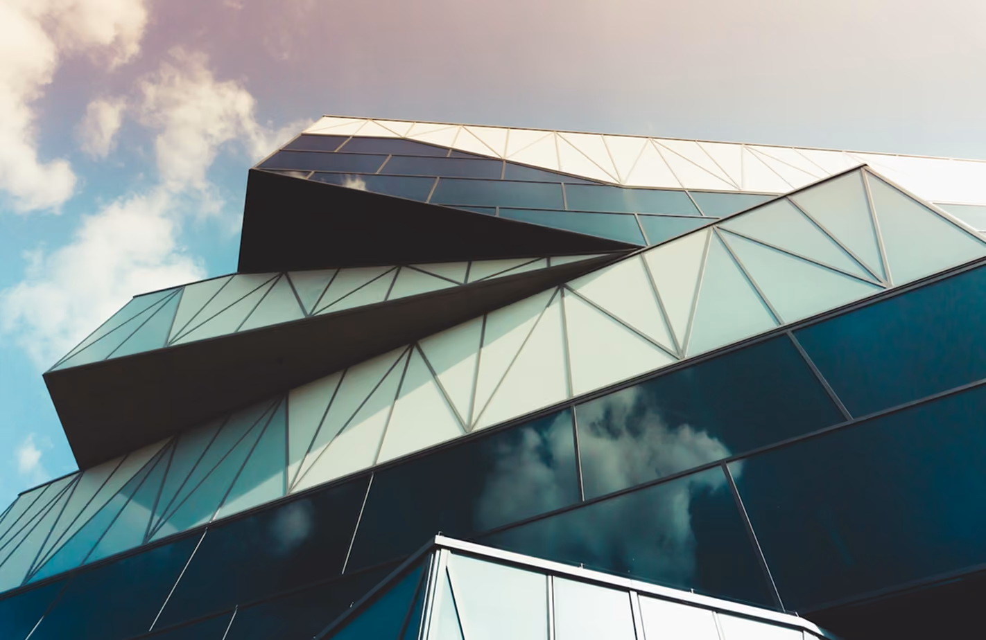 Moderne Architektur mit dreieckigen und polygonalen Glasfassadenelementen, die in einem geometrischen Muster angeordnet sind, aufgenommen aus der Froschperspektive mit Himmel und Wolken im Hintergrund.