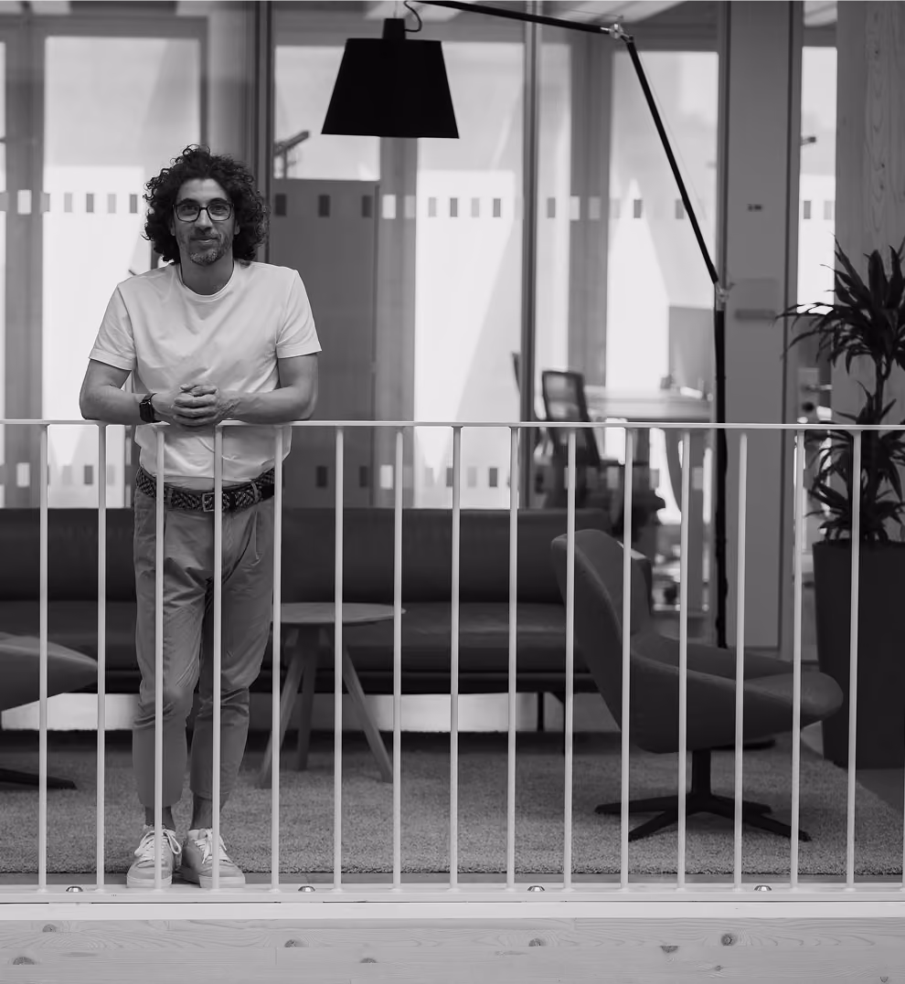 Ein Mann mit lockigem Haar, Brille, weißem T-Shirt und Stoffhose lehnt sich an ein Geländer in einem modernen Büro mit Stühlen, Sofas und einer großen Stehlampe.