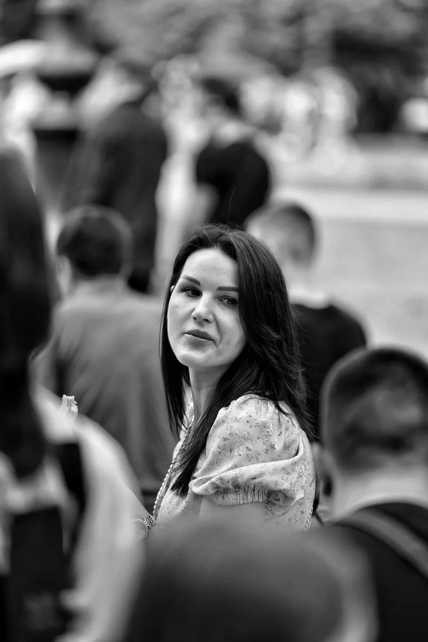Black and white portrait of a woman with dark hair looking to the side in a crowded outdoor setting.